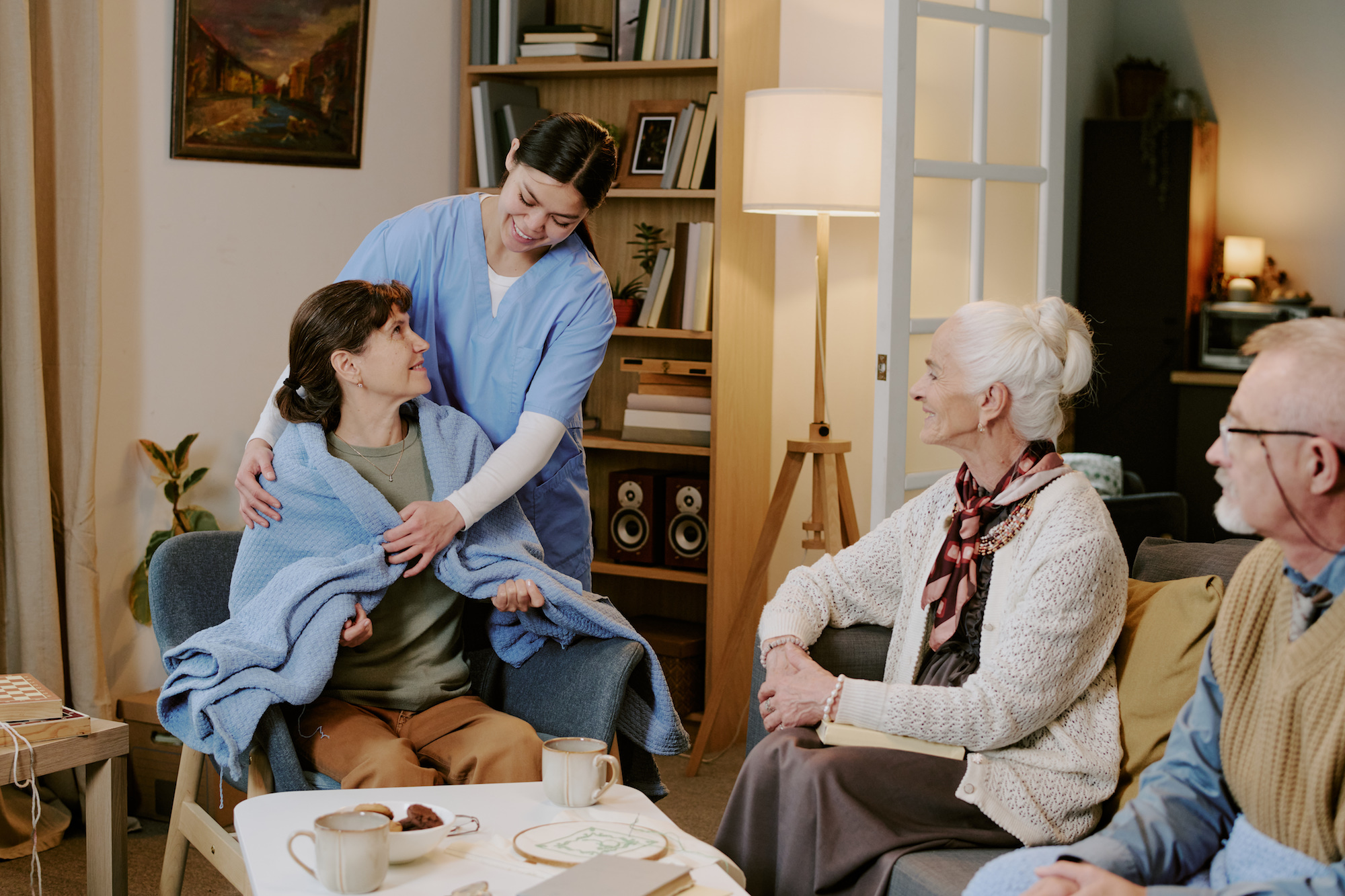 Young adult Caucasian female nurse assisting middle aged Caucasian woman with blanket while senior Caucasian woman and senior Caucasian man sitting nearby observing interaction